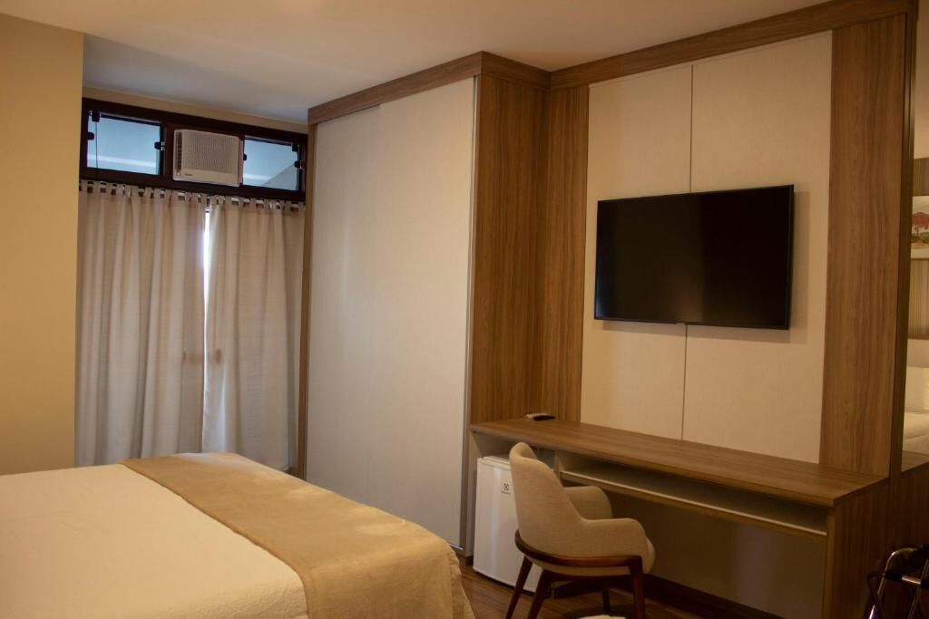 A hotel room with a bed, a desk with a chair, a television mounted on the wall, and a window with curtains.