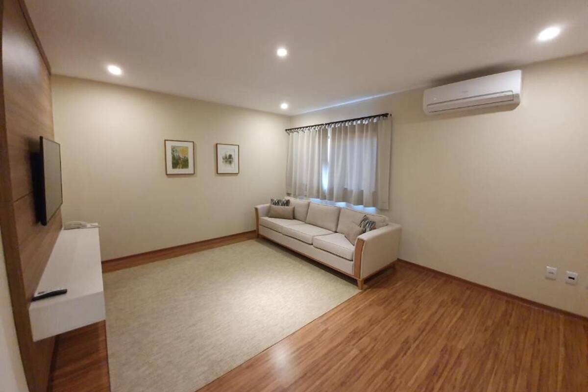 A living room with a sofa, a TV, a window with curtains, and an air conditioning unit.
