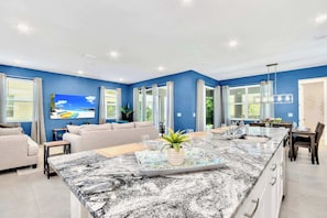 A modern kitchen with a marble countertop, a dining area with a table and chairs, and a living area with a sofa and a TV.