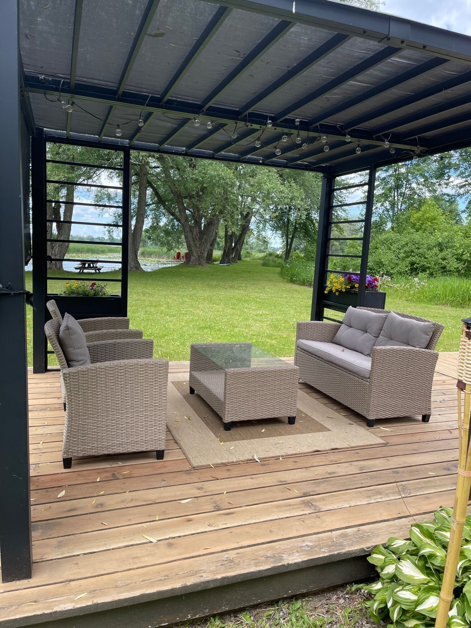 An outdoor patio with wicker furniture, a glass-top table, and a covered area with a view of a lake and trees.