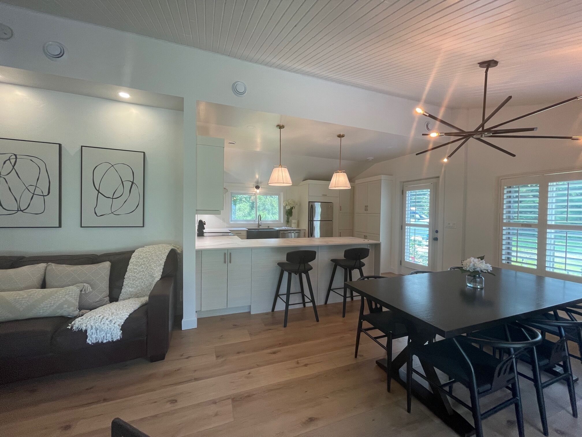 Une cuisine moderne avec des meubles blancs, un îlot central doté de tabourets, un éclairage suspendu et une table à manger avec des chaises.