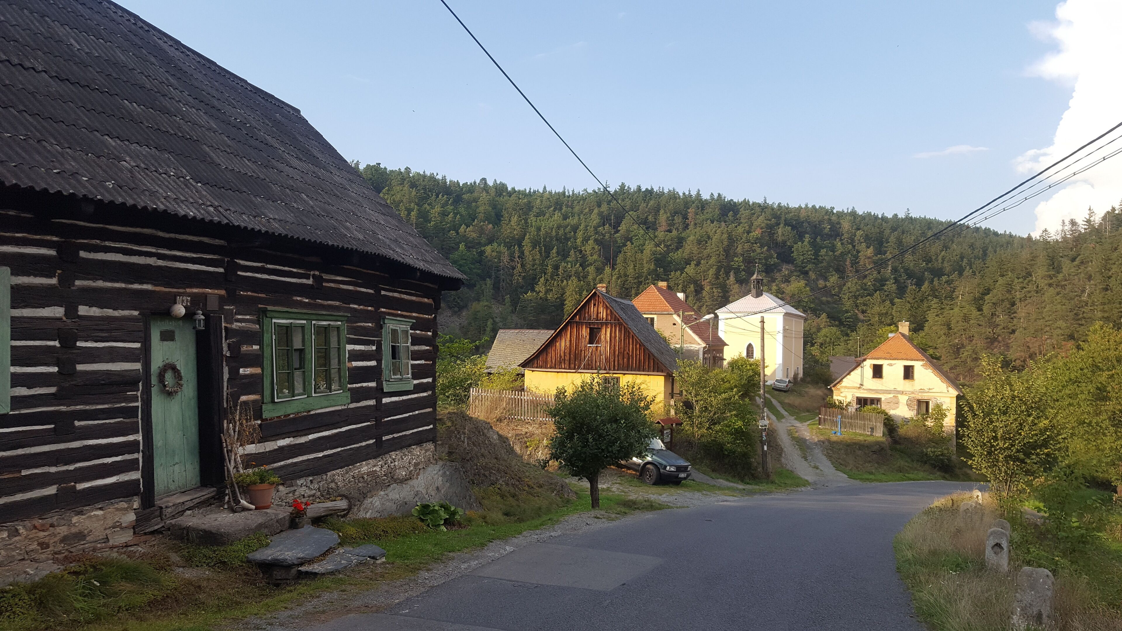 Une maison de campagne en bois, avec une porte et des fenêtres vertes, une grange jaune et une église, le tout situé dans un village.