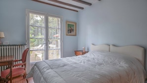 A bedroom with a large bed, a chair, a table, and a window with a view of the outdoors. - House between pine trees and beach (Saint-Brevin-les-Pins)