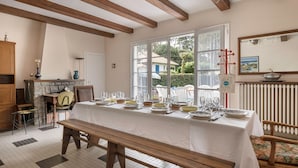 A dining area with a long table set for a meal, wooden beams on the ceiling, a window with a view of greenery, and a radiator on the wall. - House between pine trees and beach (Saint-Brevin-les-Pins)