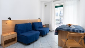 Habitación pequeña con cabecera de madera, dos sofás azules, una mesita con mantel a cuadros y una silla de madera.