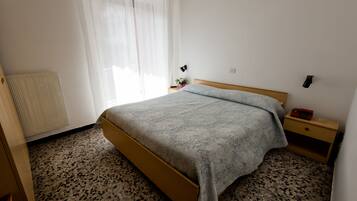 A bedroom with a bed, a bedside table, a lamp, a radiator, and a window with curtains.
