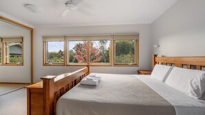 A bedroom with a wooden bed, a nightstand, a ceiling fan, and three windows offering a view of trees and a garden. - Punka River Retreat - Porepunkah, Victoria (Porepunkah)