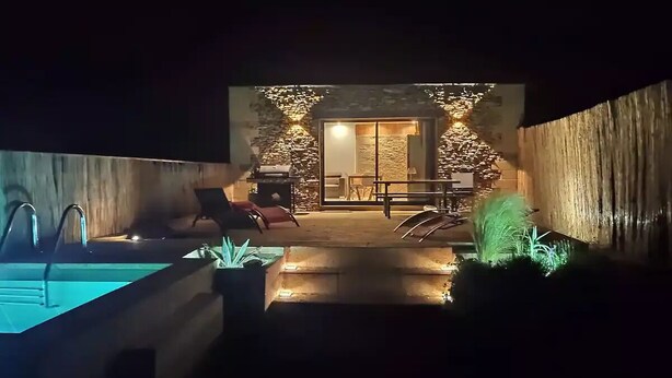 Vista nocturna de un moderno espacio exterior con piscina, una pared de piedra, una zona de descanso con sillas y una entrada bien iluminada.