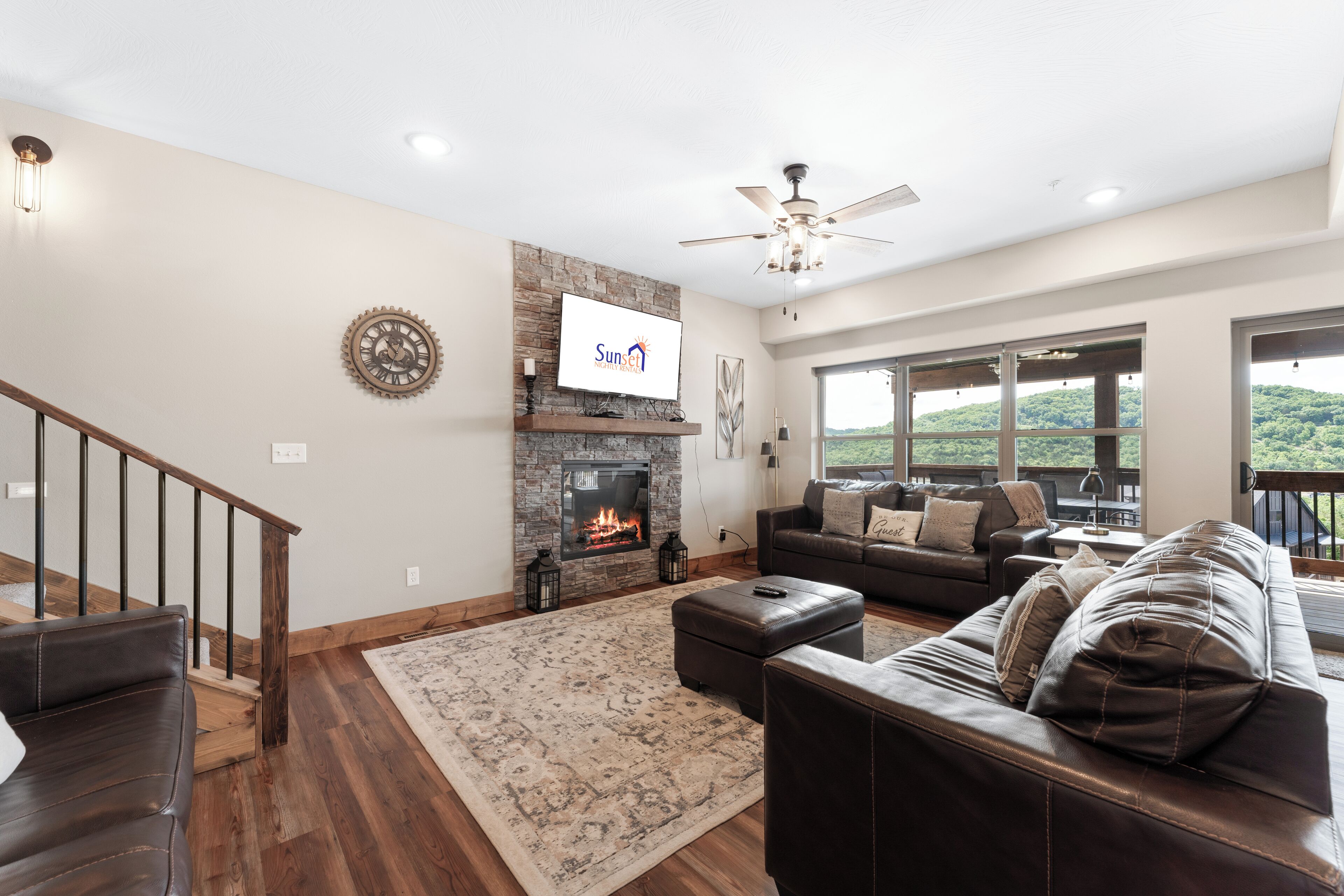 A spacious living room with a fireplace, a large window with a view, a ceiling fan, and a staircase.