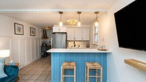 A compact kitchen with a blue island, two stools, pendant lights, and a flat-screen TV mounted on the wall.