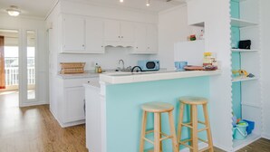 A modern kitchen with white cabinets, a teal accent wall, a microwave, and two wooden stools. - Beautiful 1 BR Condo Overlooking Freeman Park (Carolina Beach)