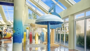 A water feature with a blue umbrella and water jets, surrounded by colorful pillars and a glass ceiling. - Dunes Village by Elliott Beach Rentals (Myrtle Beach)
