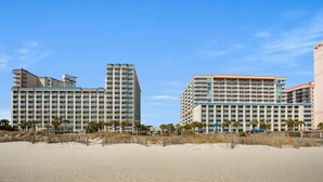 A beachfront hotel with multiple high-rise buildings, a sandy beach in the foreground, and palm trees. - Dunes Village by Elliott Beach Rentals (Myrtle Beach)