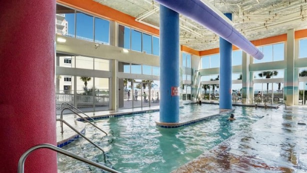 An indoor swimming pool with clear water, surrounded by large windows and blue pillars. - Dunes Village by Elliott Beach Rentals (Myrtle Beach)