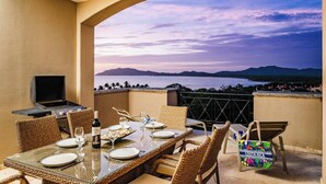 A dining area with a table set for two, a grill, and a view of a body of water and mountains. - Matapalo 602, romantic, luxurious, 2 BR apt. with stunning views (Tamarindo)