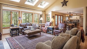 A spacious living room with a large window overlooking a forest, a cozy fireplace, and a dining area with a chandelier.