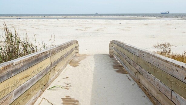 Ein hölzerner Steg führt zu einem Sandstrand, in der Ferne ein Schiff.