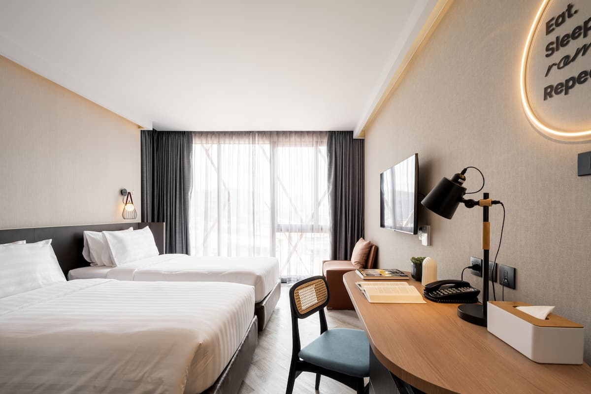 A modern hotel room with two beds, a desk with a lamp, a chair, and a wall-mounted TV.
