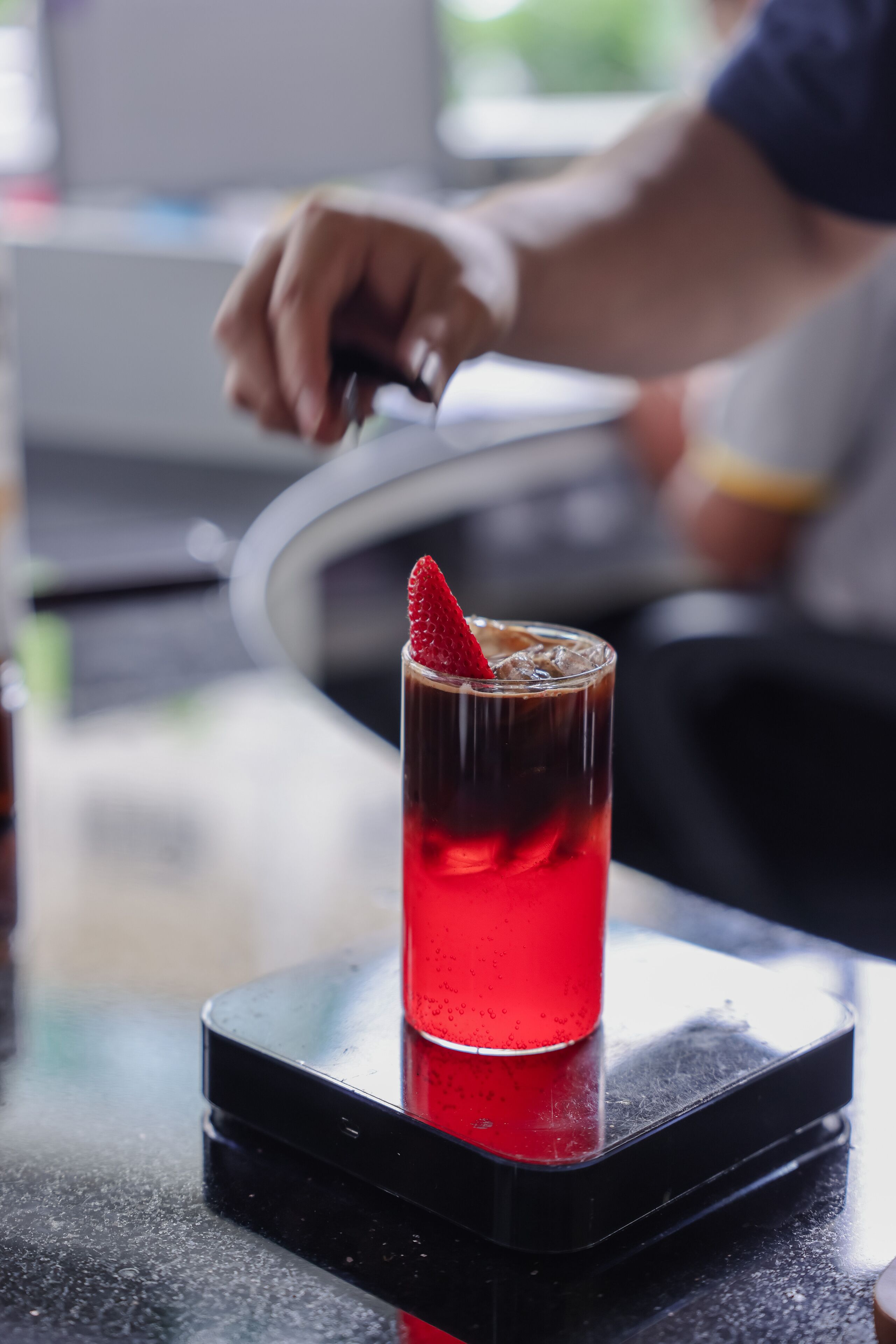 Een glas rode drank met een aardbeien garnering op een zwarte onderzetter.