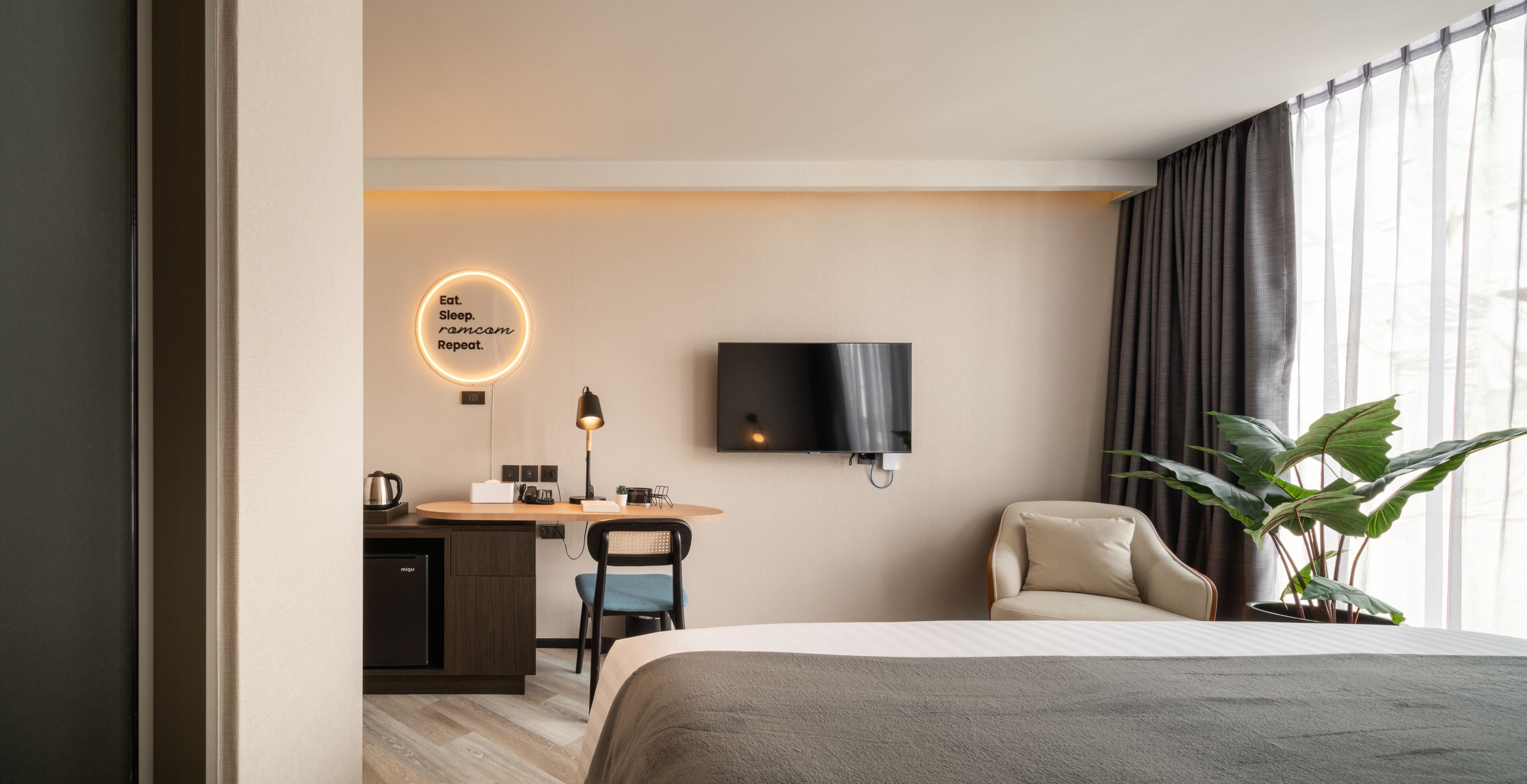 A modern hotel room with a bed, desk, chair, TV, and a plant.