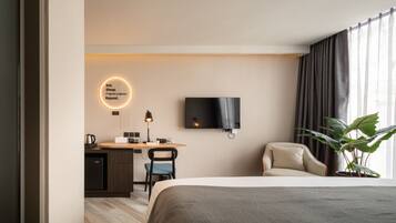 A modern hotel room with a bed, desk, chair, TV, and a plant.