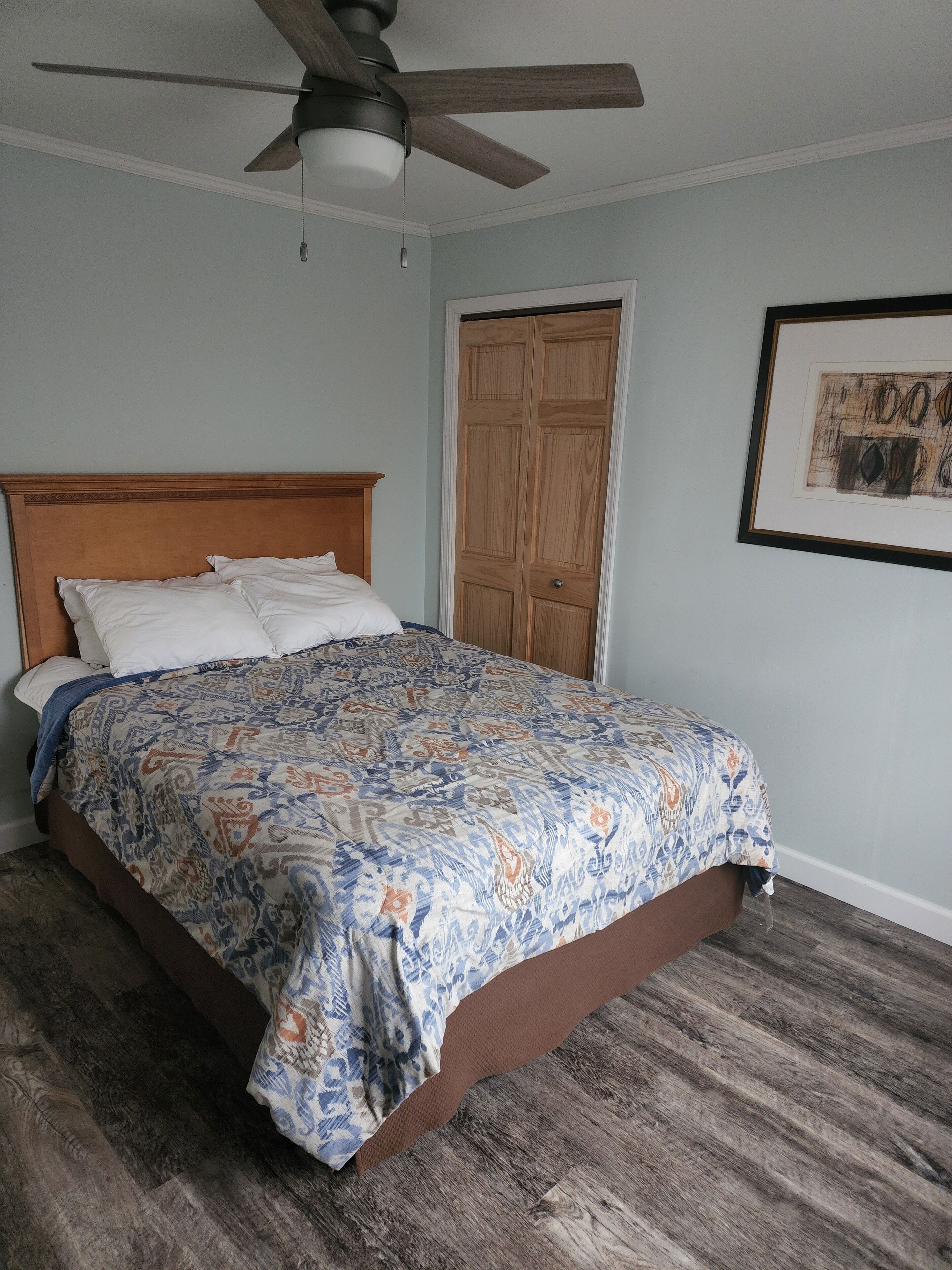 Un dormitorio con cama de madera, ventilador de techo, armario de madera y un cuadro en la pared.