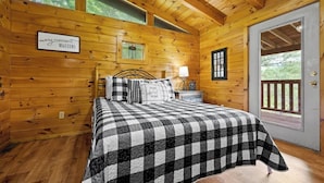 A wooden cabin bedroom with a checkered bedspread, a nightstand with a lamp, and a window with a view of trees.