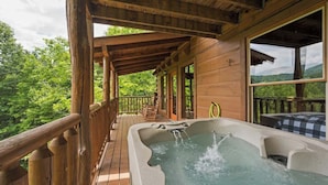 A wooden deck with a hot tub, wooden railings, and a view of a forested landscape.