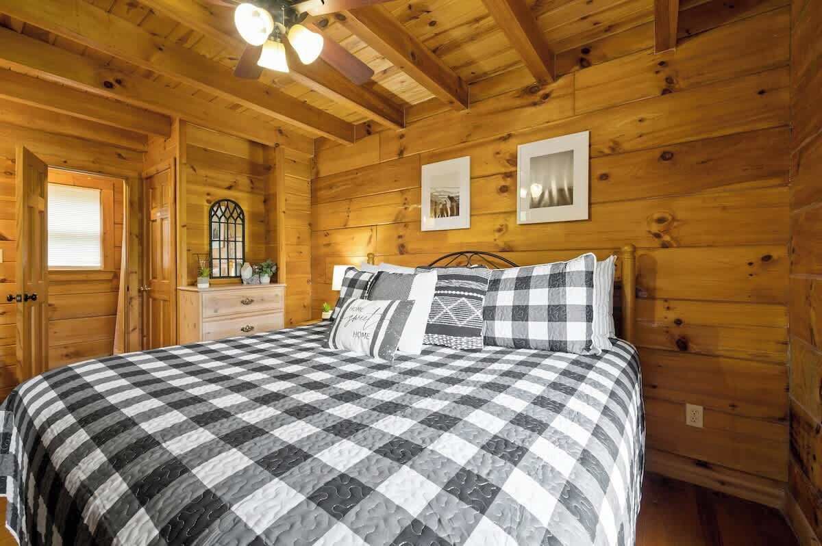 A cozy wooden cabin bedroom with a checkered bedspread, a dresser, a mirror, and framed pictures on the wall.
