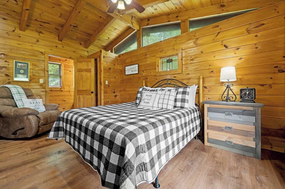 A cozy cabin room with a bed, a chair, a dresser, and a ceiling fan.