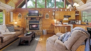 A cozy cabin interior with a stone fireplace, wooden walls, a kitchen area, and a comfortable seating area.
