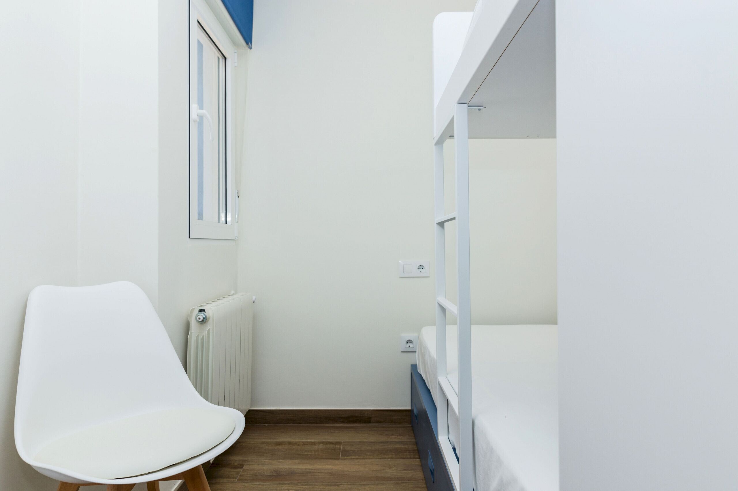 Una habitación estrecha con una silla blanca, camas literas, un radiador y una ventana.