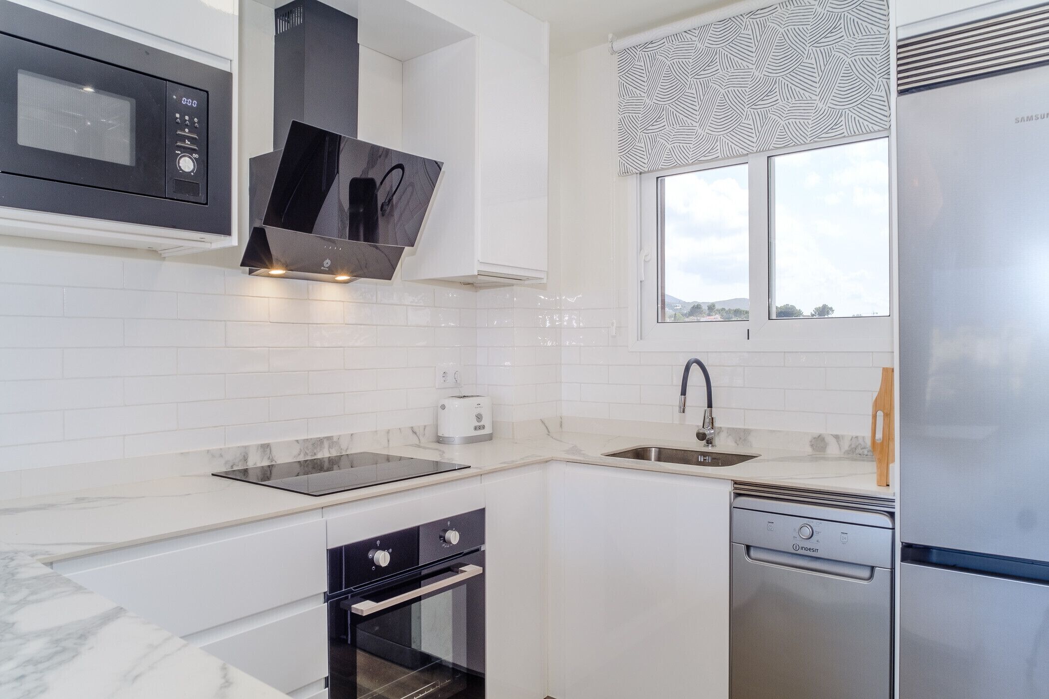 A modern kitchen with white cabinetry, a built-in microwave, a stainless steel dishwasher, and a window with patterned curtains.