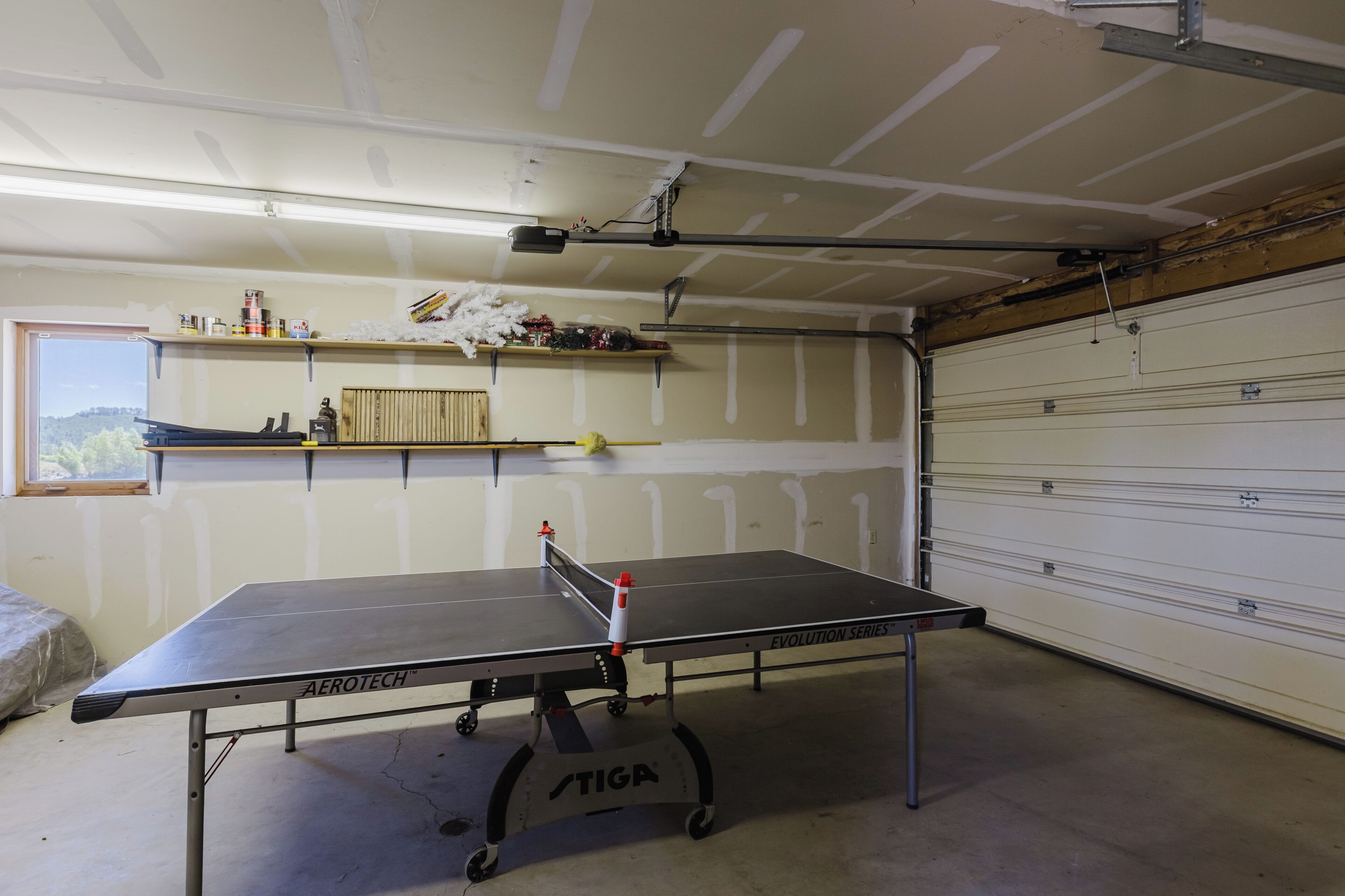 A ping pong table in a garage with a wooden ceiling, a window, and a garage door.
