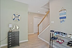 A hallway with a staircase, a life preserver decoration, a dresser with drawers, and a shoe rack with various pairs of shoes.