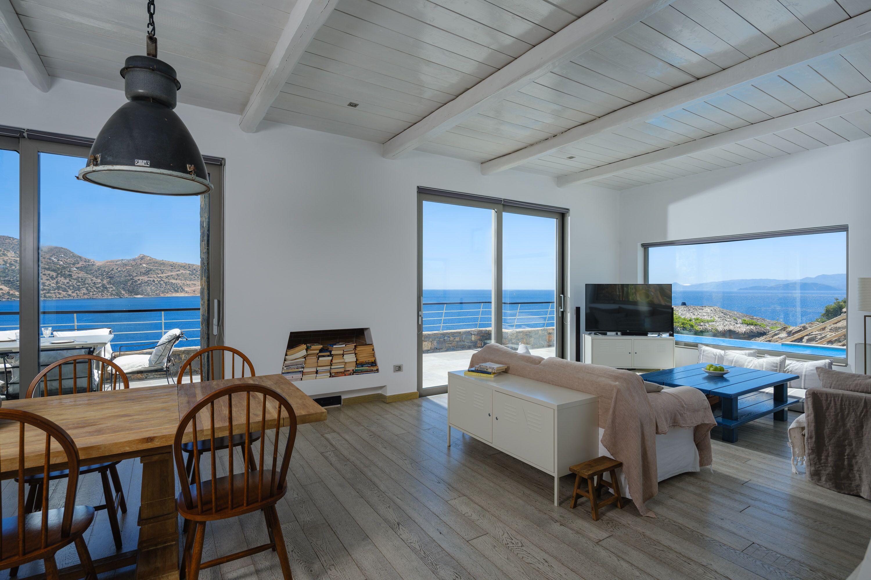 Un vaste espace de vie comprenant une table à manger en bois, des chaises, un canapé et un téléviseur. De grandes fenêtres offrent une vue sur la mer et les montagnes.
