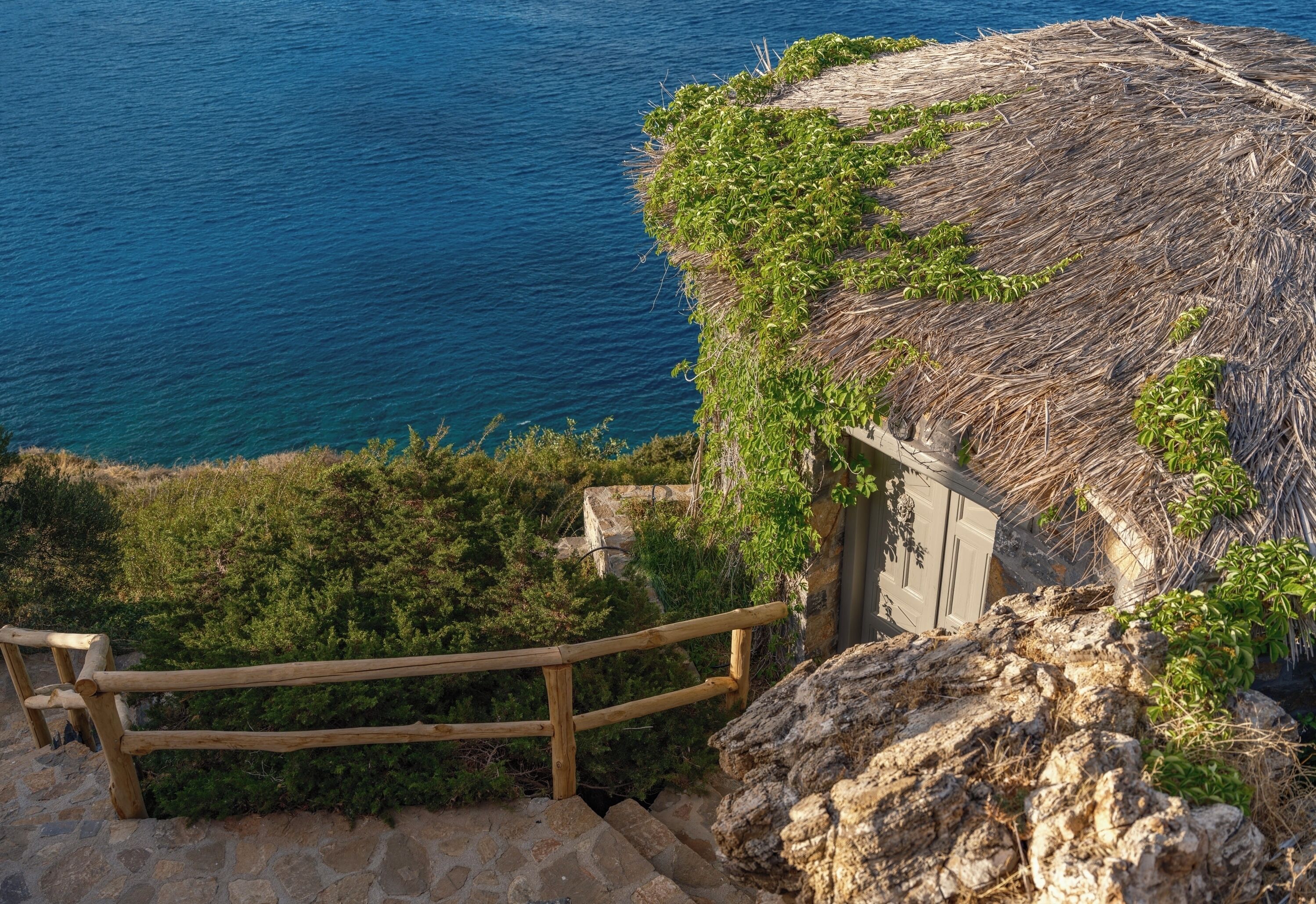 Une construction rustique à toit de chaume, dotée d’un escalier en bois menant à l’entrée, entourée de verdure et offrant une vue sur une étendue d’eau.