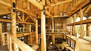 A spacious log cabin interior with a central seating area, wooden beams, and large windows.