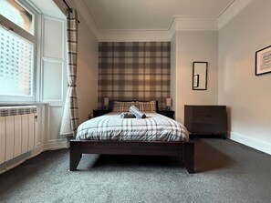 A bedroom with a bed, a nightstand, a dresser, a radiator, and a window with curtains. - Belford Hostel (Edinburgh)