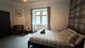 A bedroom with a bed, a chair, a small table, a window with curtains, and a radiator.