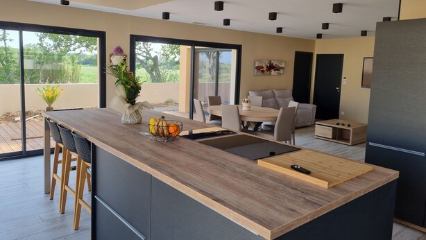 A modern kitchen with a wooden island, a dining area with a sofa, and a large window offering a view of the outdoors. - La Villa Manon - FLORENSAC (Florensac)