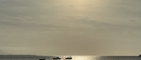 A beach scene with the sun setting over the water, boats, and a person in the water.