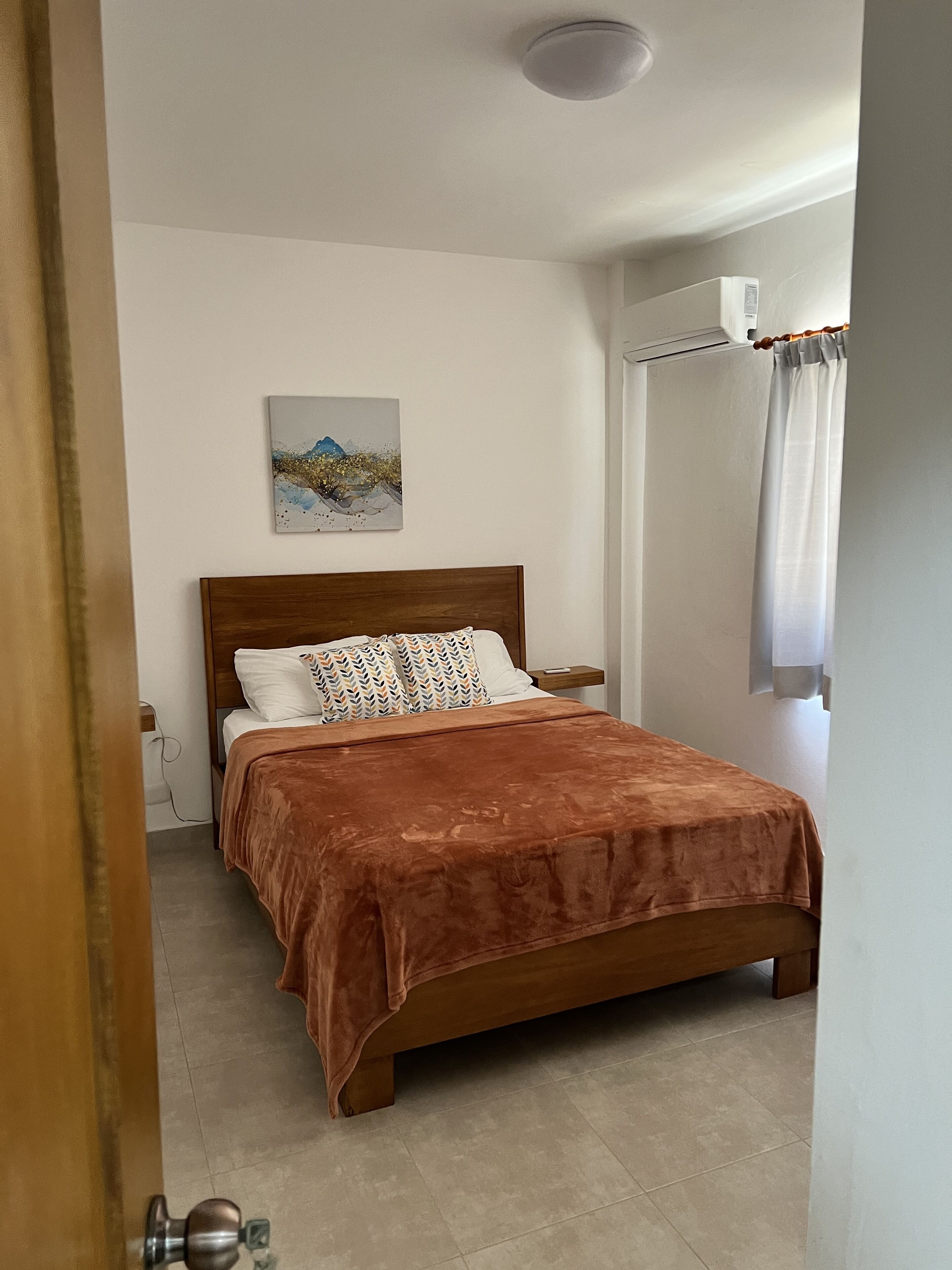 Une chambre à coucher avec un lit en bois, un mur blanc, un tableau et une fenêtre avec des rideaux.