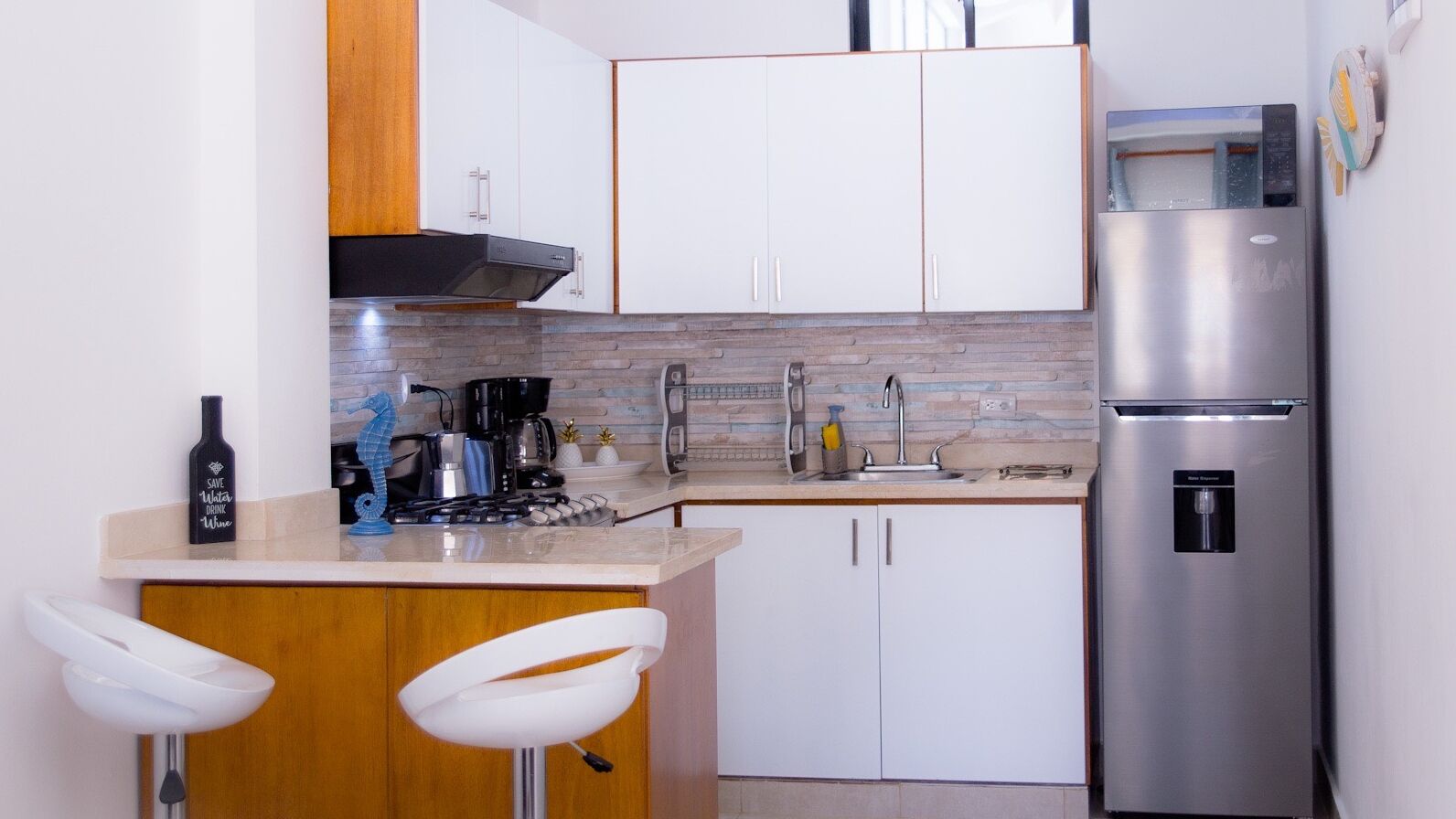 Una cocina compacta con armarios blancos, un refrigerador de acero inoxidable, una cafetera y dos taburetes de bar blancos.