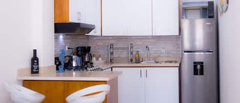 Una cocina compacta con armarios blancos, un refrigerador de acero inoxidable, una cafetera y dos taburetes de bar blancos.