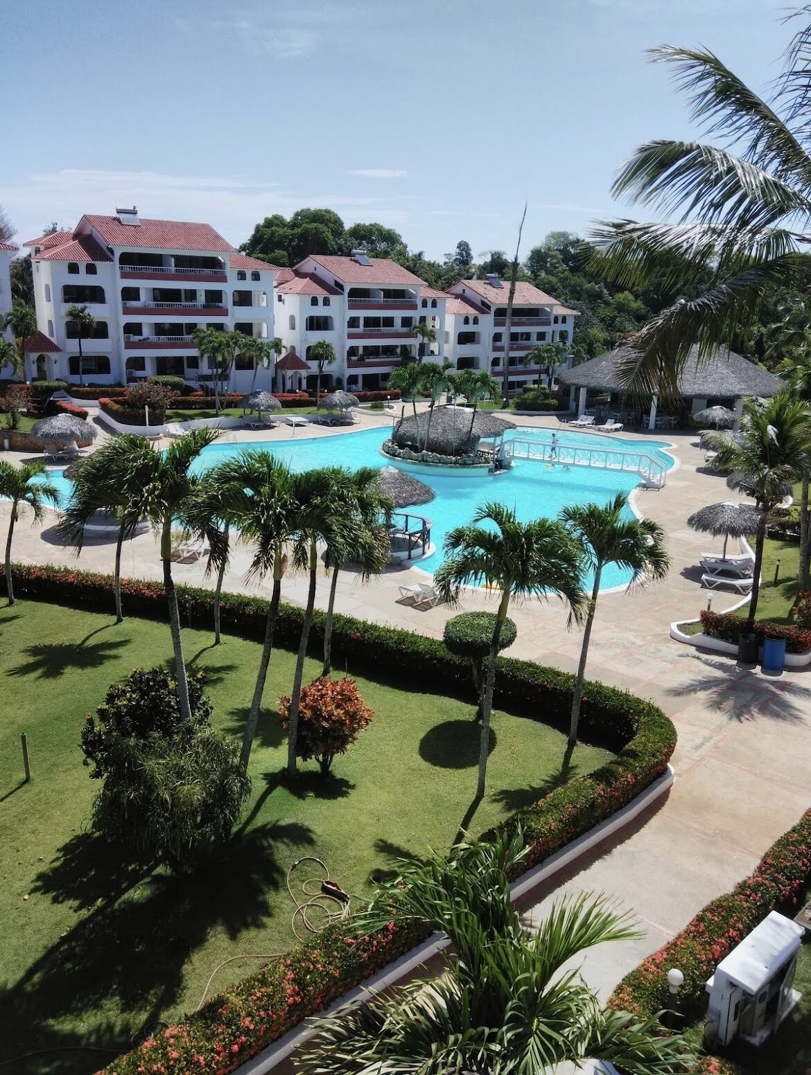 A resort with a large central pool, palm trees, and a multi-story building with balconies.