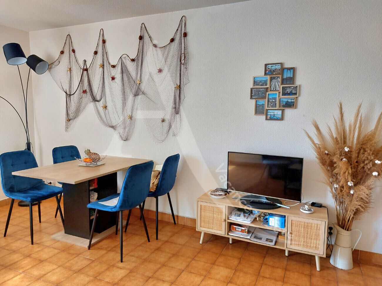 A modern living room with a wooden dining table, blue chairs, a flat-screen TV, a cabinet with electronic devices, and decorative wall art.