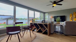 A modern living room with a large window, a ceiling fan, a desk with a laptop, a grey sofa, and a TV.