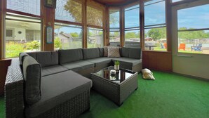 A sunroom with a large gray sectional sofa, a glass coffee table, and a view of a backyard with outdoor furniture.