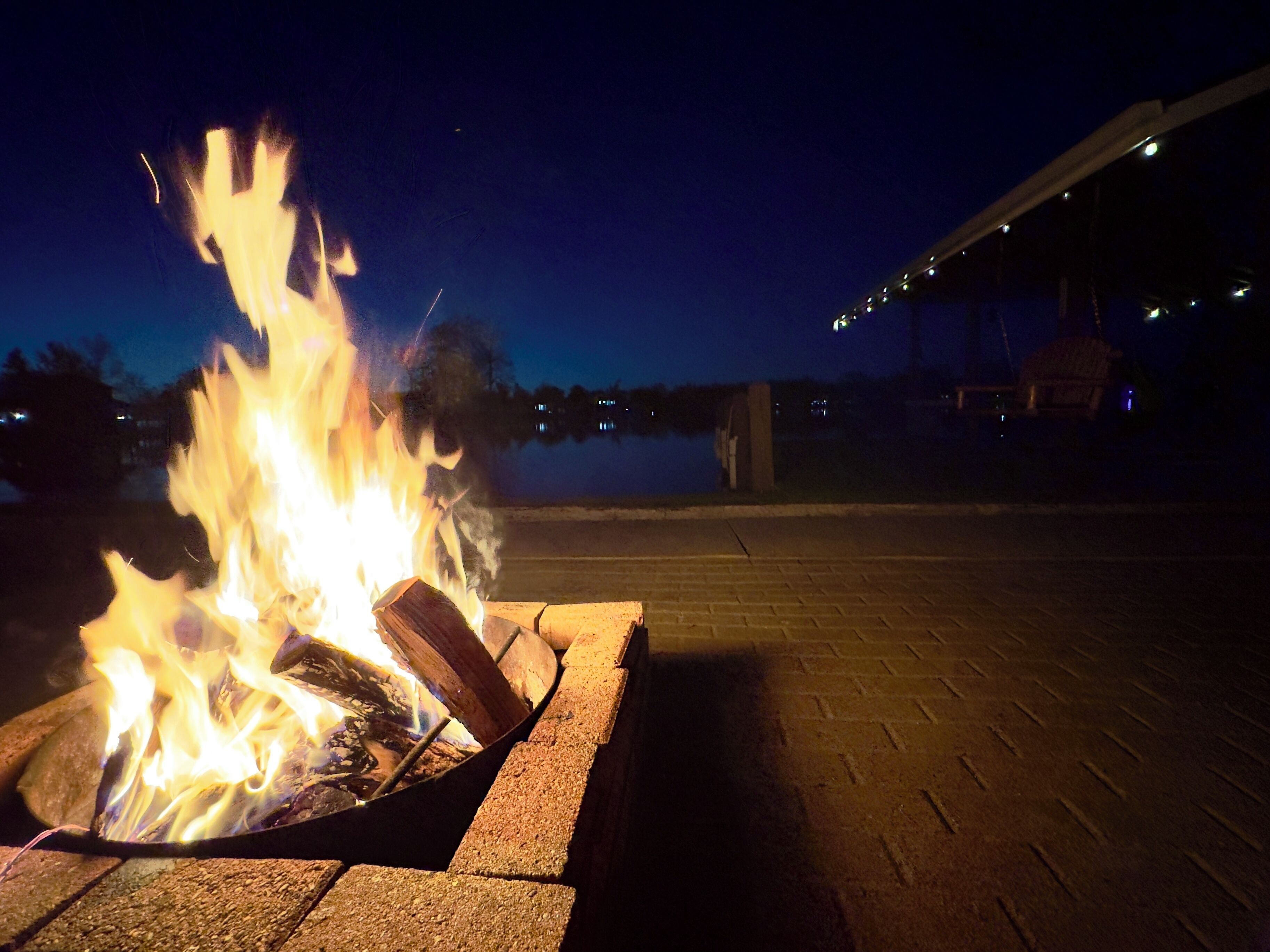 A fire pit with flames, surrounded by a brick border, set on a paved area with a body of water and a building in the background.